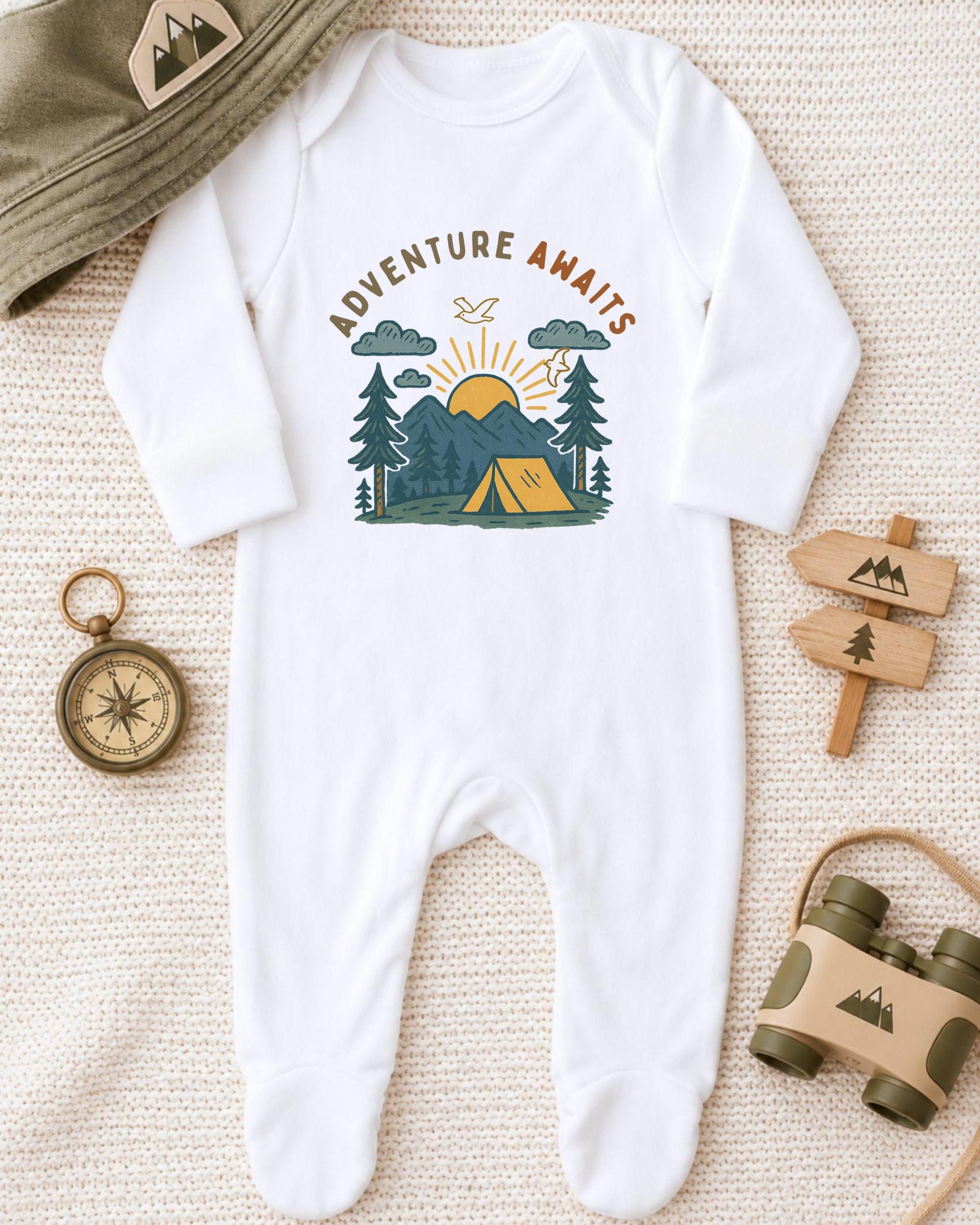A white onesie with a graphic design of a camping scene, a compass, and mountains, along with a hat and a pair of binoculars, all laid out on a beige surface.