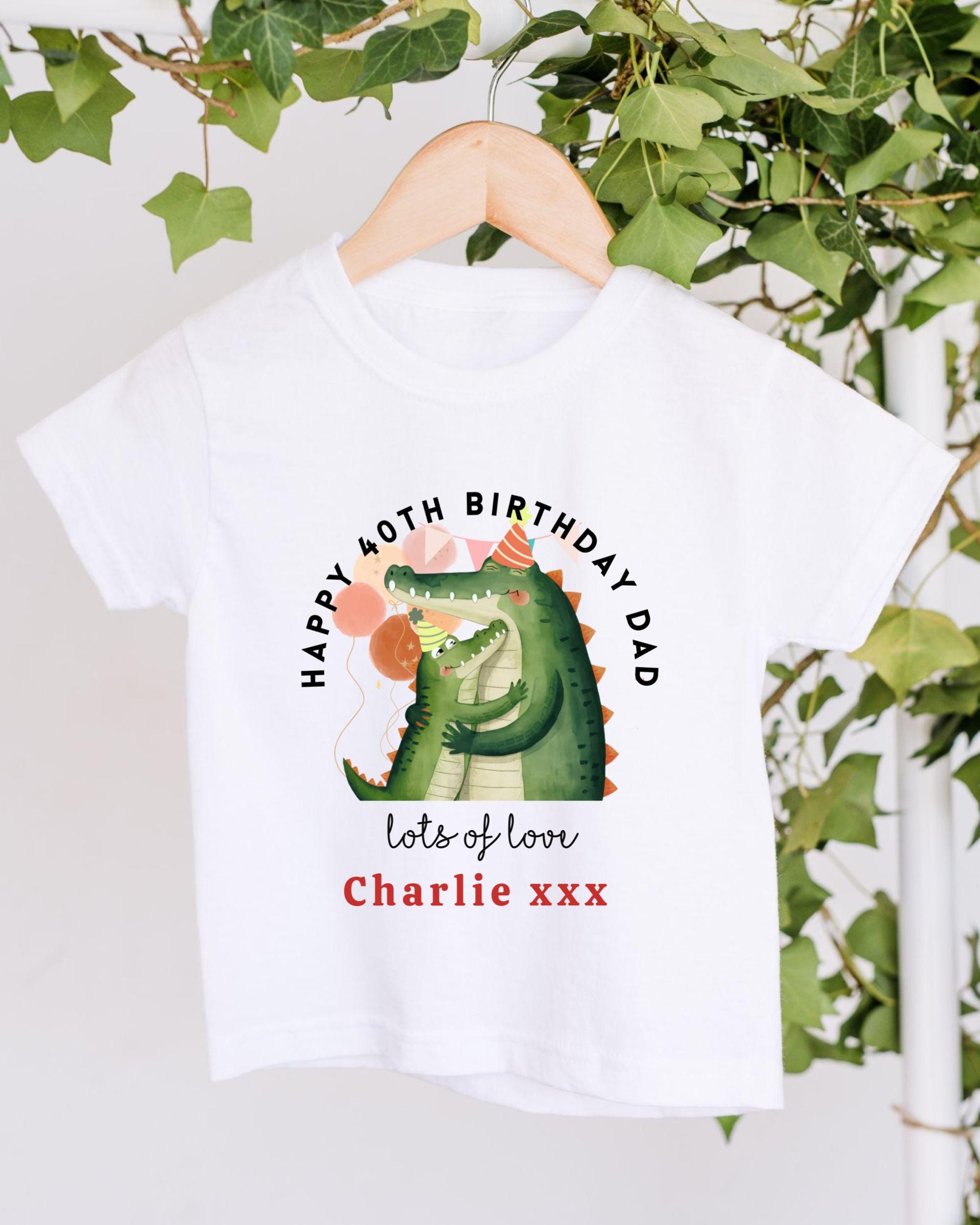A white t-shirt with a graphic design featuring a green alligator wearing a party hat and holding a balloon. The text on the t-shirt reads "Happy 40th Birthday Dad lots of love Charlie xxx".