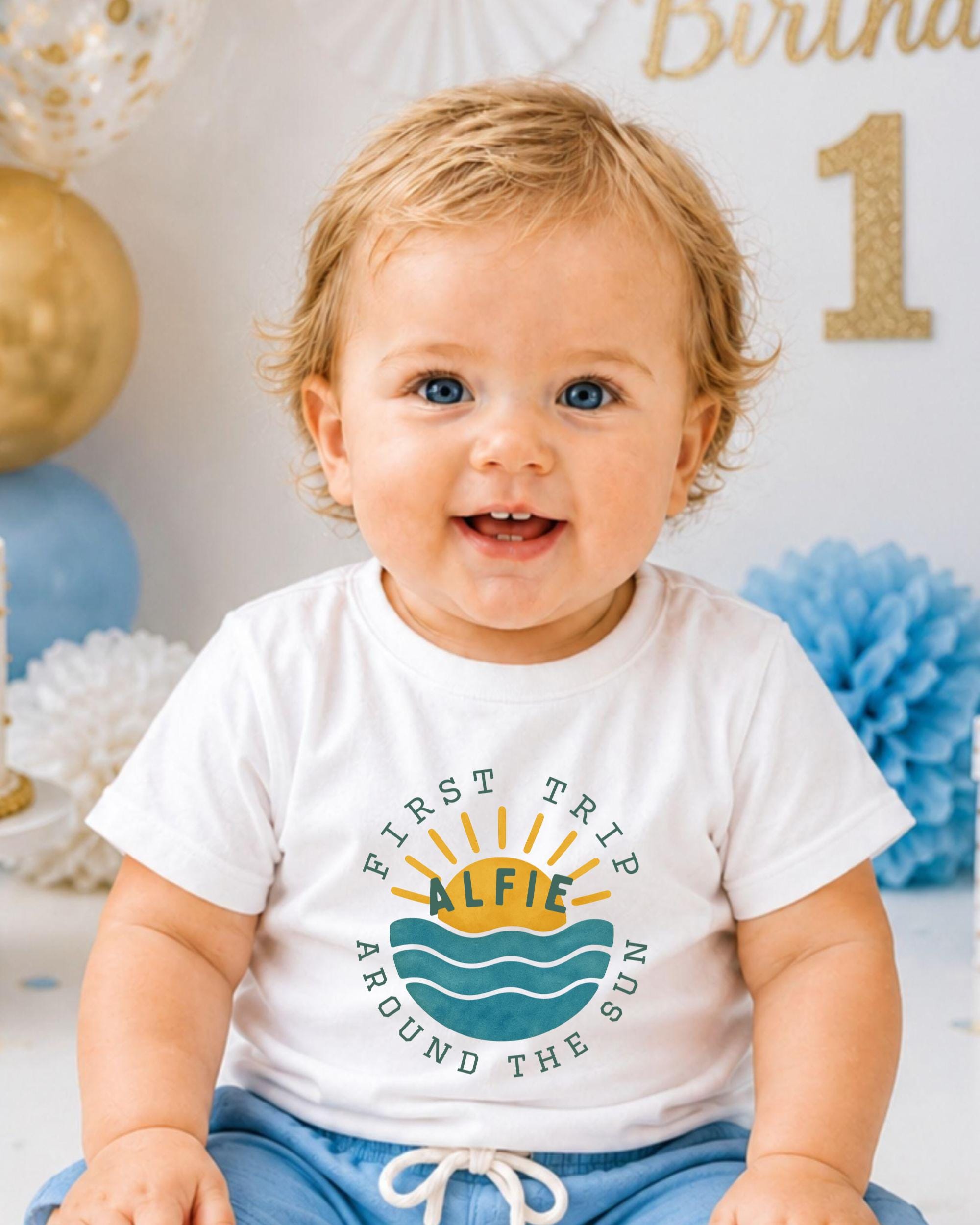 A smiling baby boy wearing a white t-shirt with a graphic design and text, sitting in front of a birthday party backdrop with balloons and a number 1.