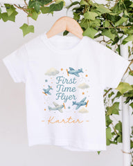 A white t-shirt with a graphic design featuring airplanes and clouds, along with the text "First Time Flyer" and the name "Kater" printed on it.
