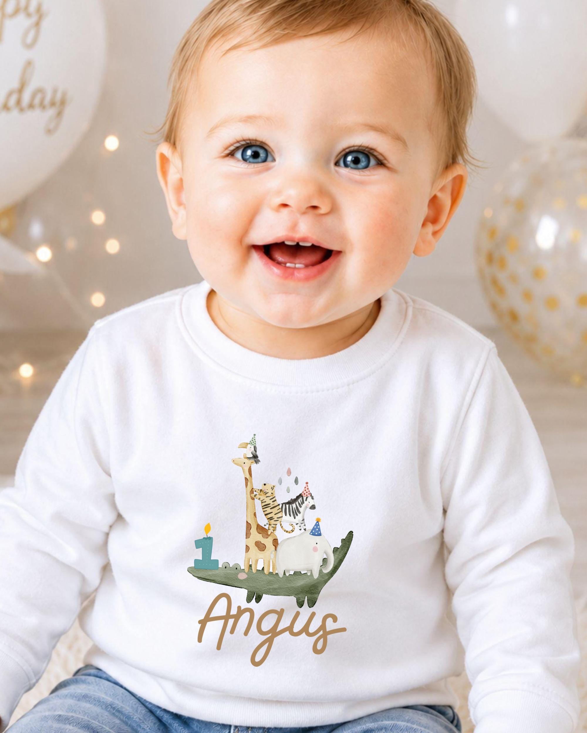 A smiling baby boy wearing a white shirt with a cartoon animal design and the name "Angus" printed on it.