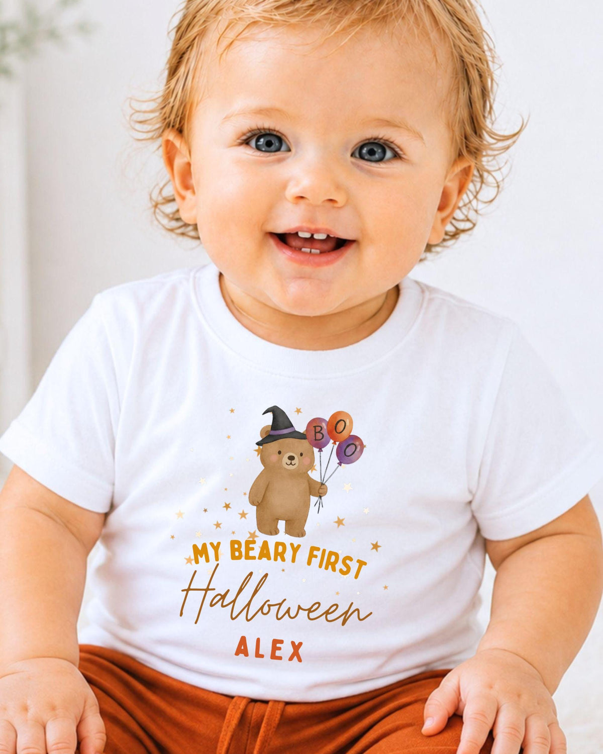 A smiling baby wearing a white t-shirt with a cartoon bear and text that says "My Beary First Halloween Alex".