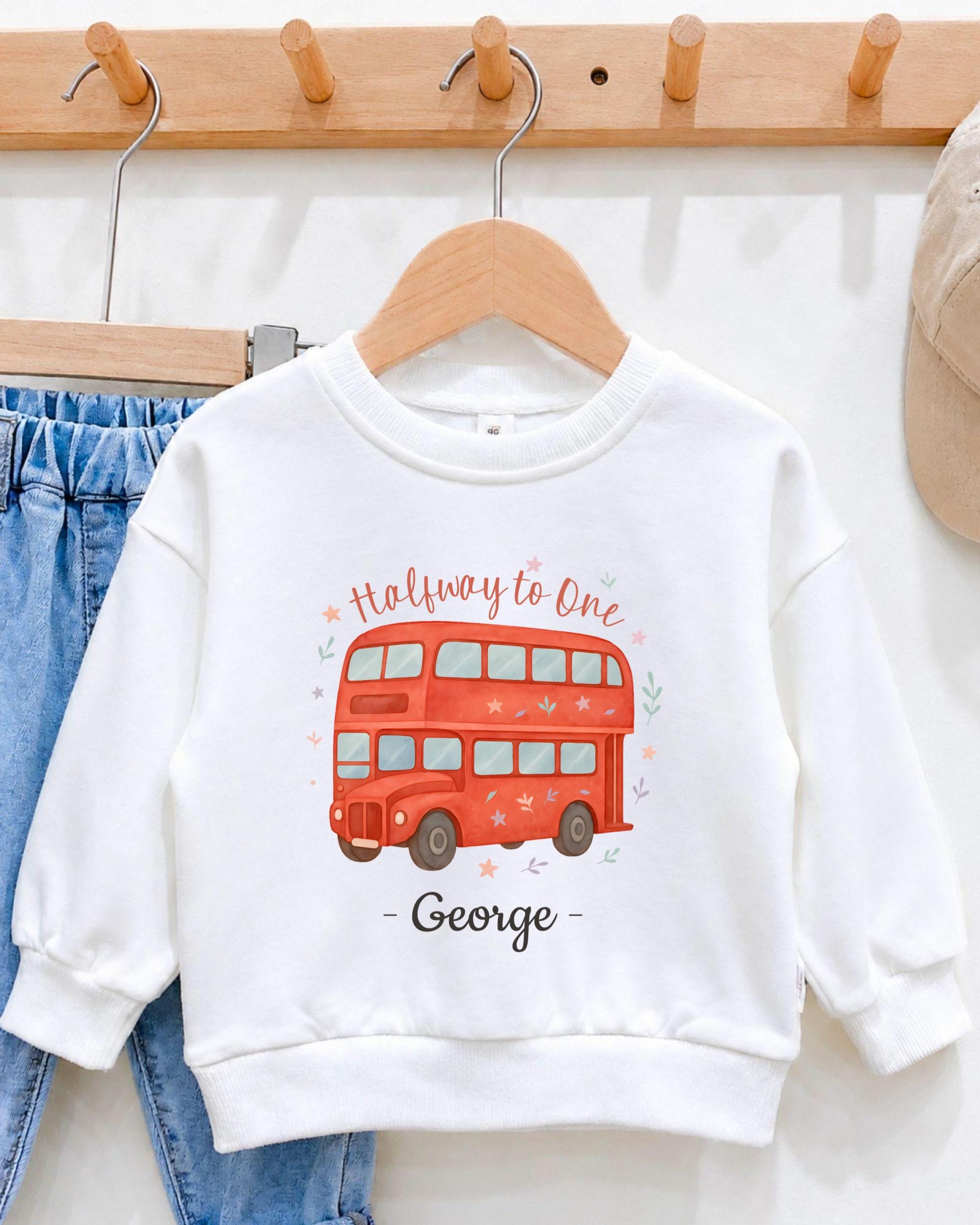 A white sweatshirt with a red double-decker bus design and the text "Halfway to One George" printed on it, hanging on a wooden hanger.