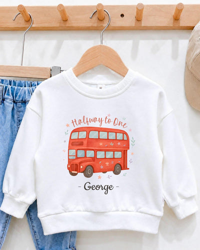 A white sweatshirt with a red double-decker bus design and the text "Halfway to One George" printed on it, hanging on a wooden hanger.