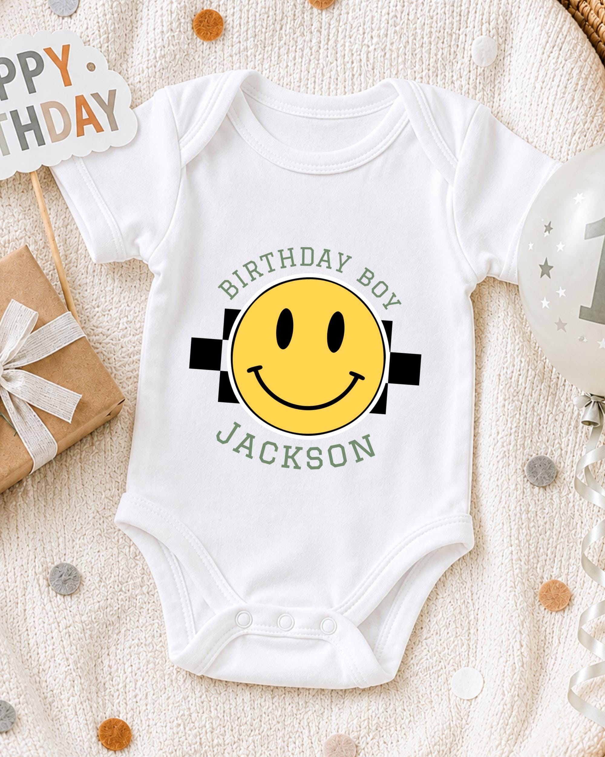 A white onesie with a yellow smiley face and the text "Birthday Boy Jackson" printed on it, along with a "Happy Birthday" sign and confetti.