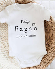 A white baby onesie with the text "Baby Fagan Coming Soon" printed on it, placed on a woven basket.