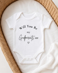 A white onesie with the text "Will You Be my Godparents xxx" printed on it, placed in a woven basket.