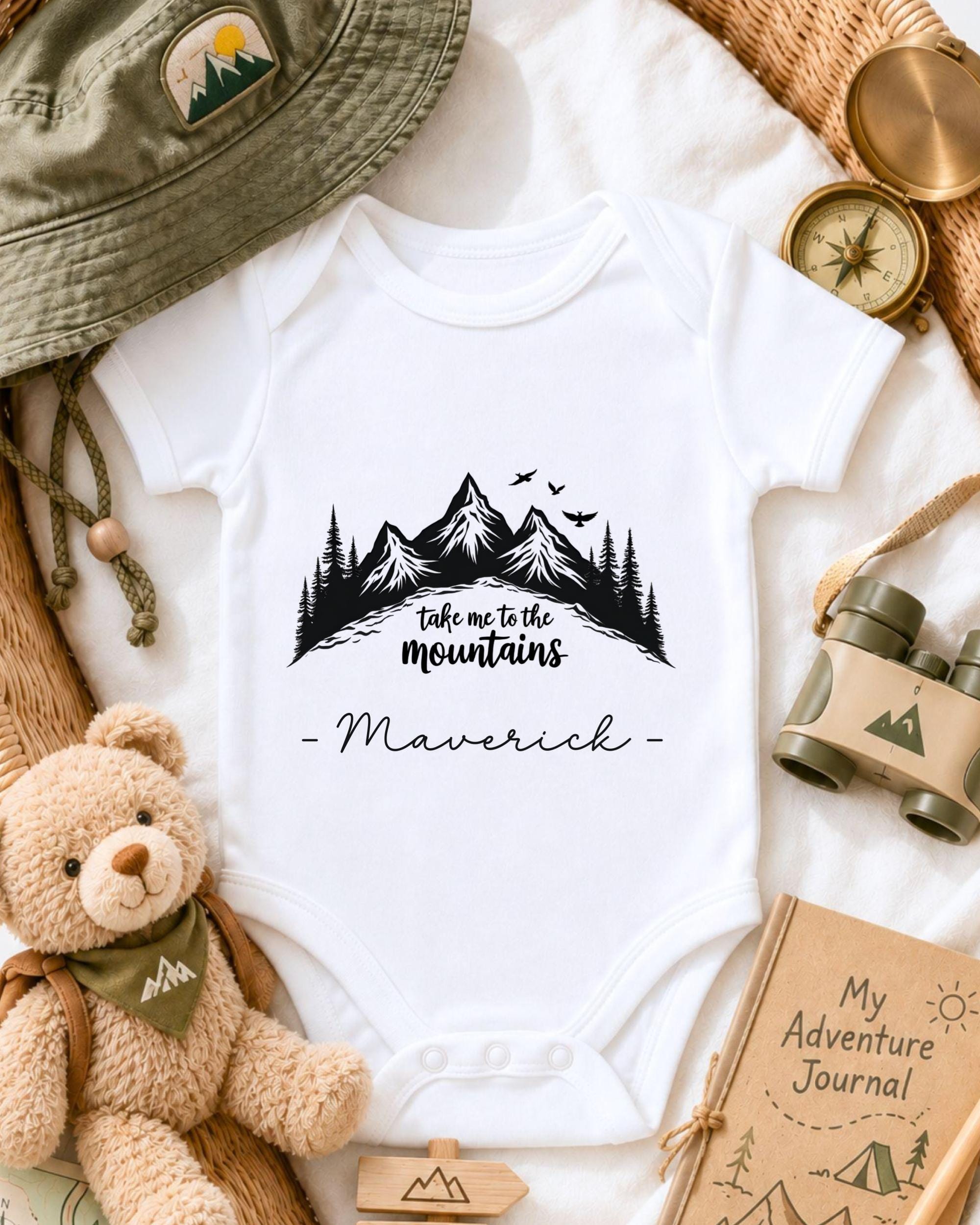 a baby onesie with a mountain landscape design, surrounded by various outdoor-themed items such as a hat, compass, binoculars, and a teddy bear.