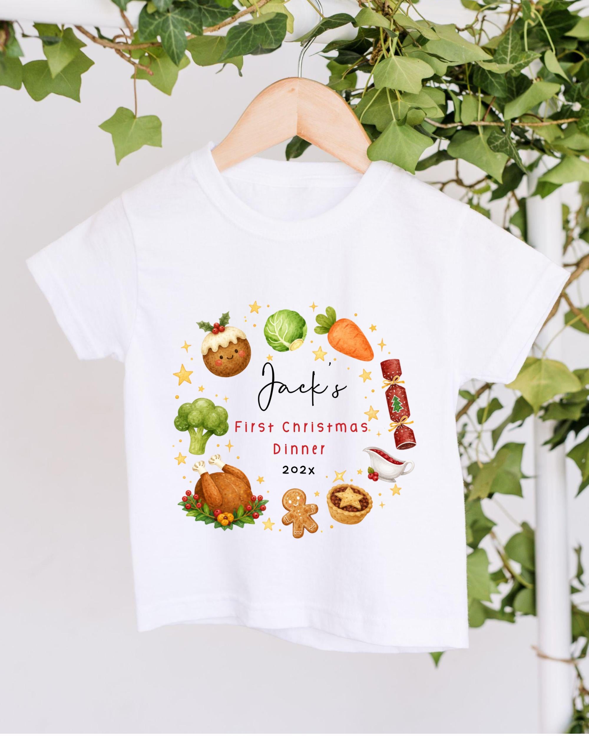 A white t-shirt with a festive design featuring various Christmas-themed items such as a carrot, broccoli, gingerbread man, and a gingerbread man. The text on the t-shirt reads "Jack's First Christmas Dinner 2023".