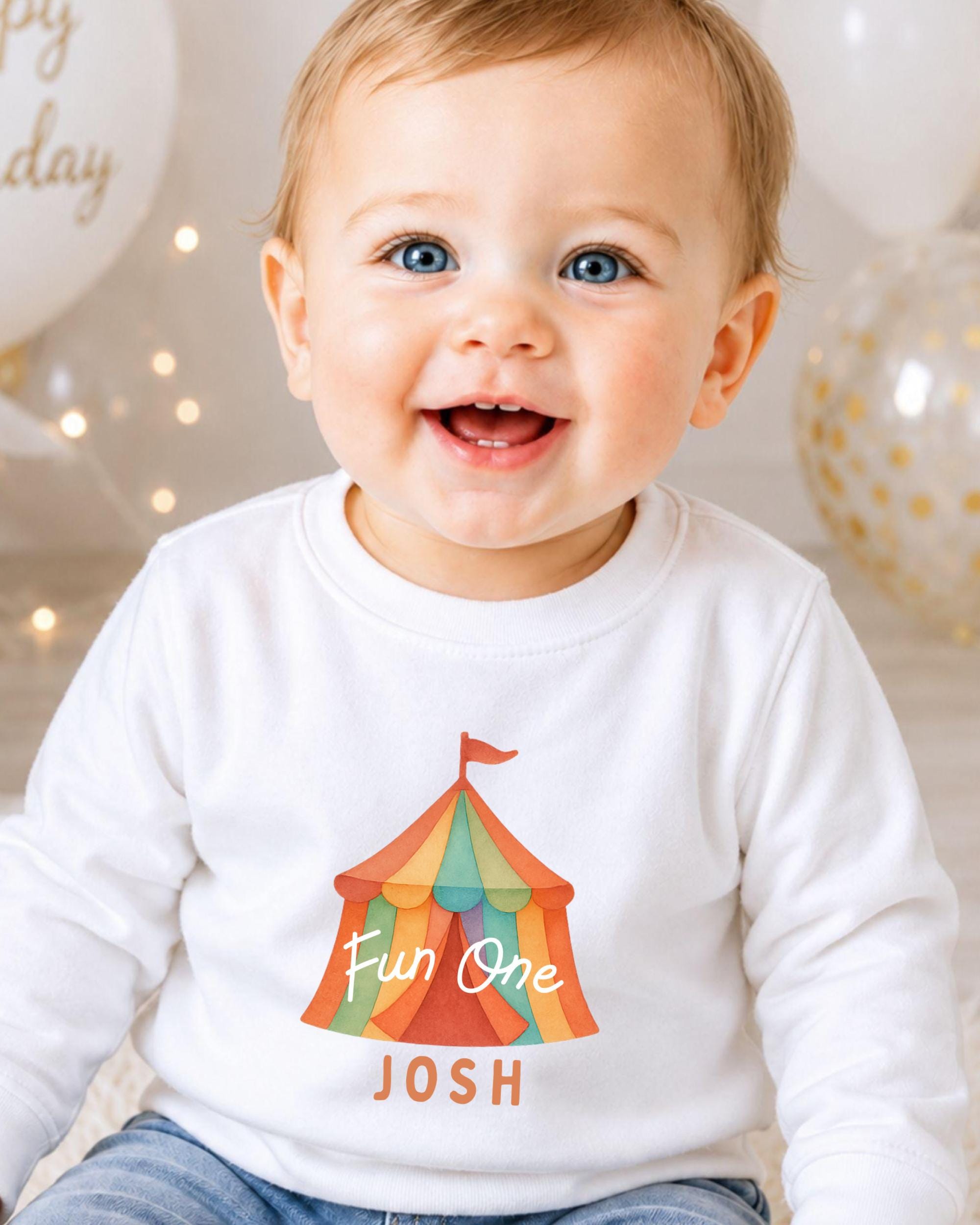 A smiling baby boy wearing a white shirt with a colorful circus tent graphic and the name " Josh" printed on it.