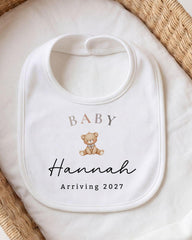 A white baby bib with a teddy bear and the text "BALY Hannah Arriving 2027" printed on it.