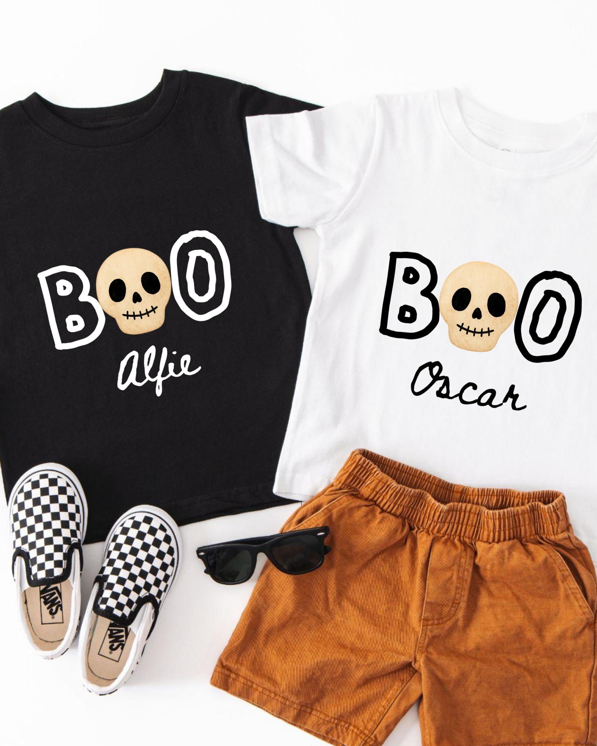 A black t-shirt with a white skull design and the text "BOO Alfred" printed on it, a white t-shirt with the same text "BOO Oscar", a pair of brown shorts, and a pair of black and white checkered sneakers.