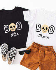 A black t-shirt with a white skull design and the text "BOO Alfred" printed on it, a white t-shirt with the same text "BOO Oscar", a pair of brown shorts, and a pair of black and white checkered sneakers.