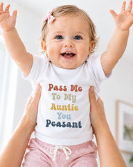 A smiling baby girl wearing a white t-shirt with text and a pink bow, being held up by someone's arms.