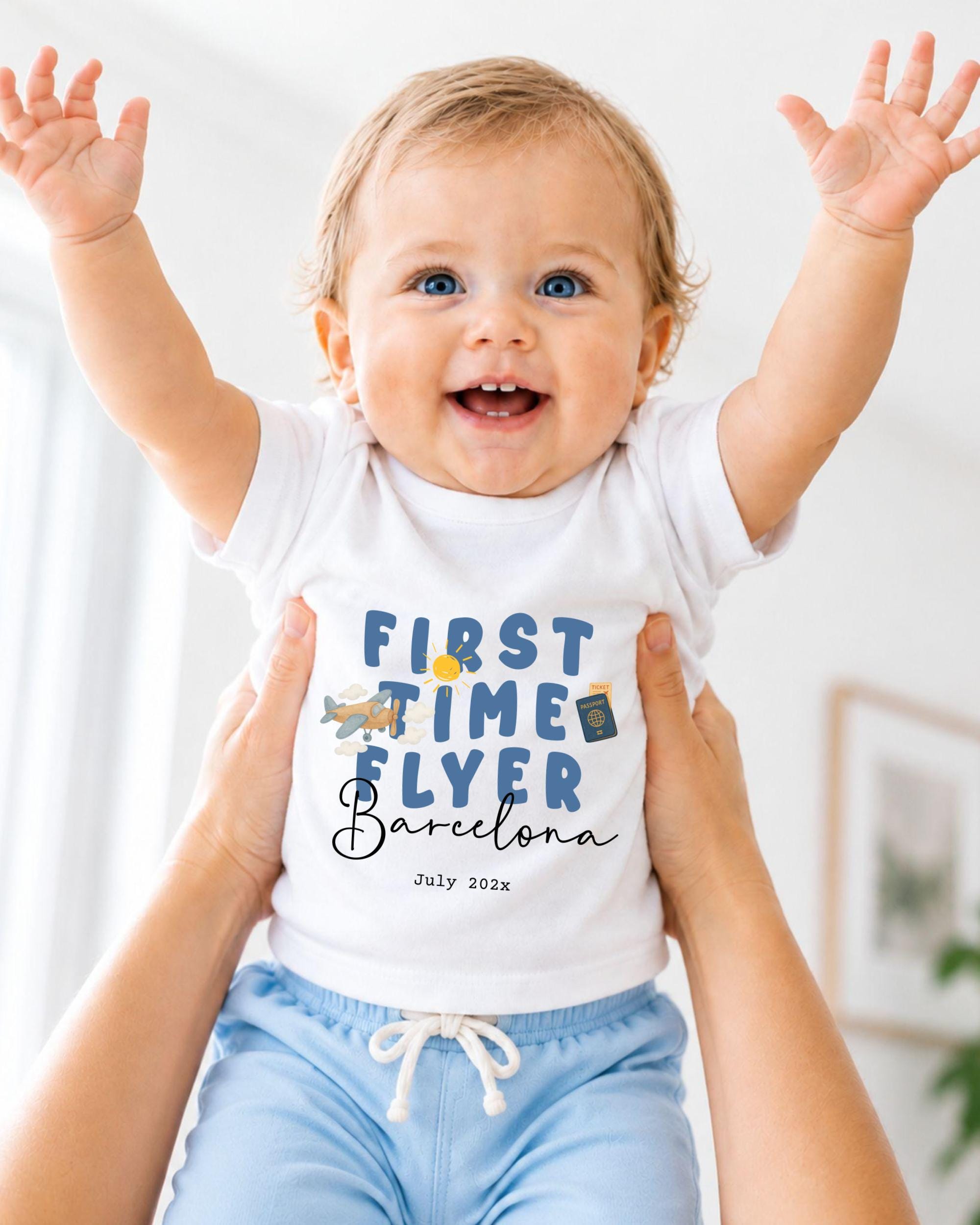 A smiling baby wearing a white t-shirt with the text "FIRST TIME FLYER Barcelona" and blue shorts, being held up by someone's arms.