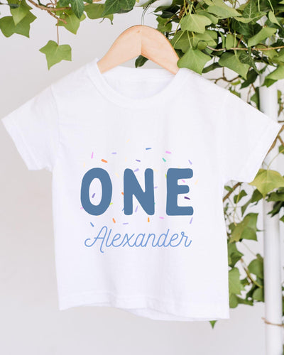 A white t-shirt with the text "ONE Alexander" printed on it, along with colorful sprinkles. The t-shirt is hanging on a wooden hanger, with a plant visible in the background.