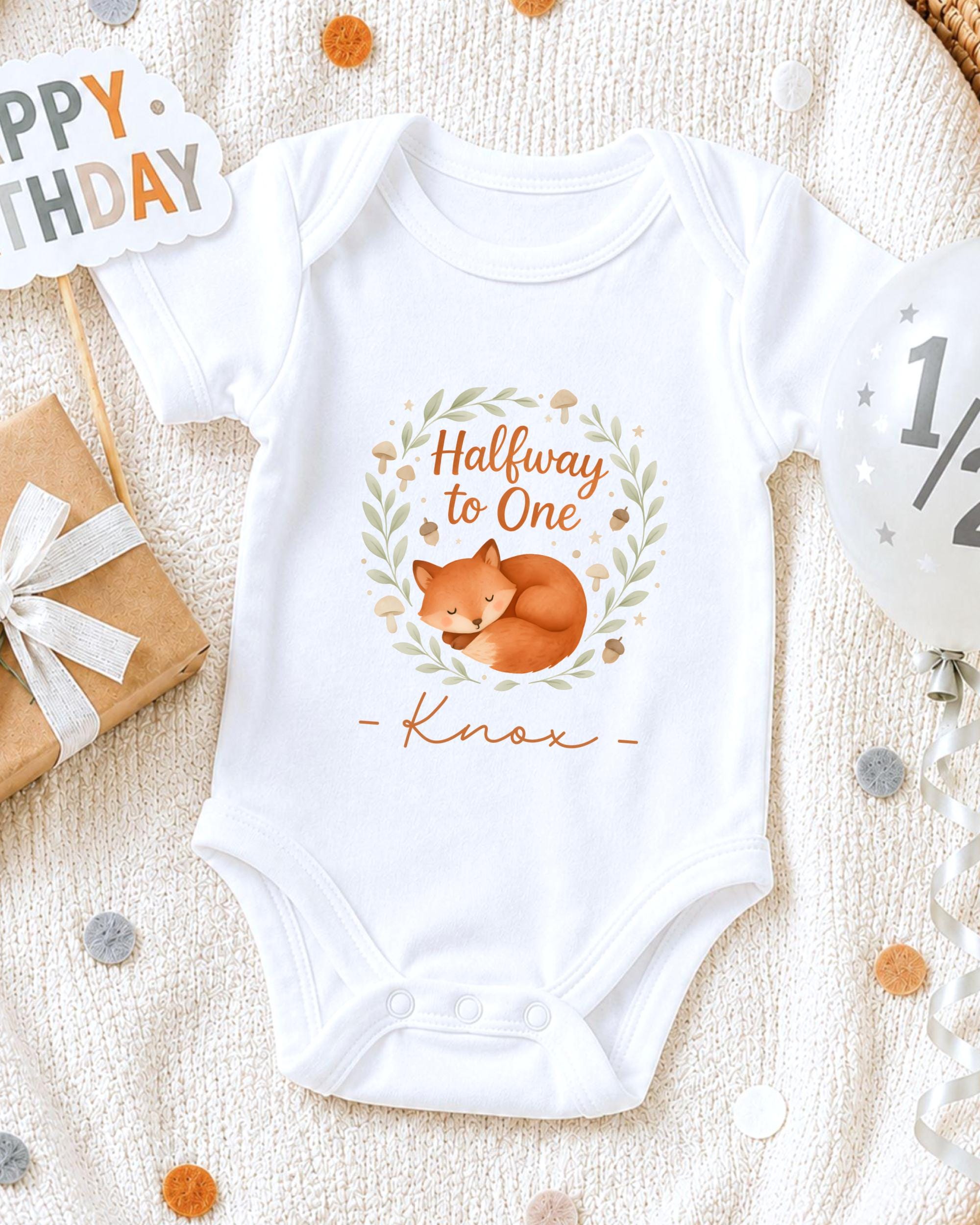 A white onesie with a cartoon image of a fox and the text "Halfway to One" printed on it, along with the name "Knox" below the image. The onesie is placed on a white blanket, surrounded by various party decorations such as a birthday sign, a balloon, and confetti.