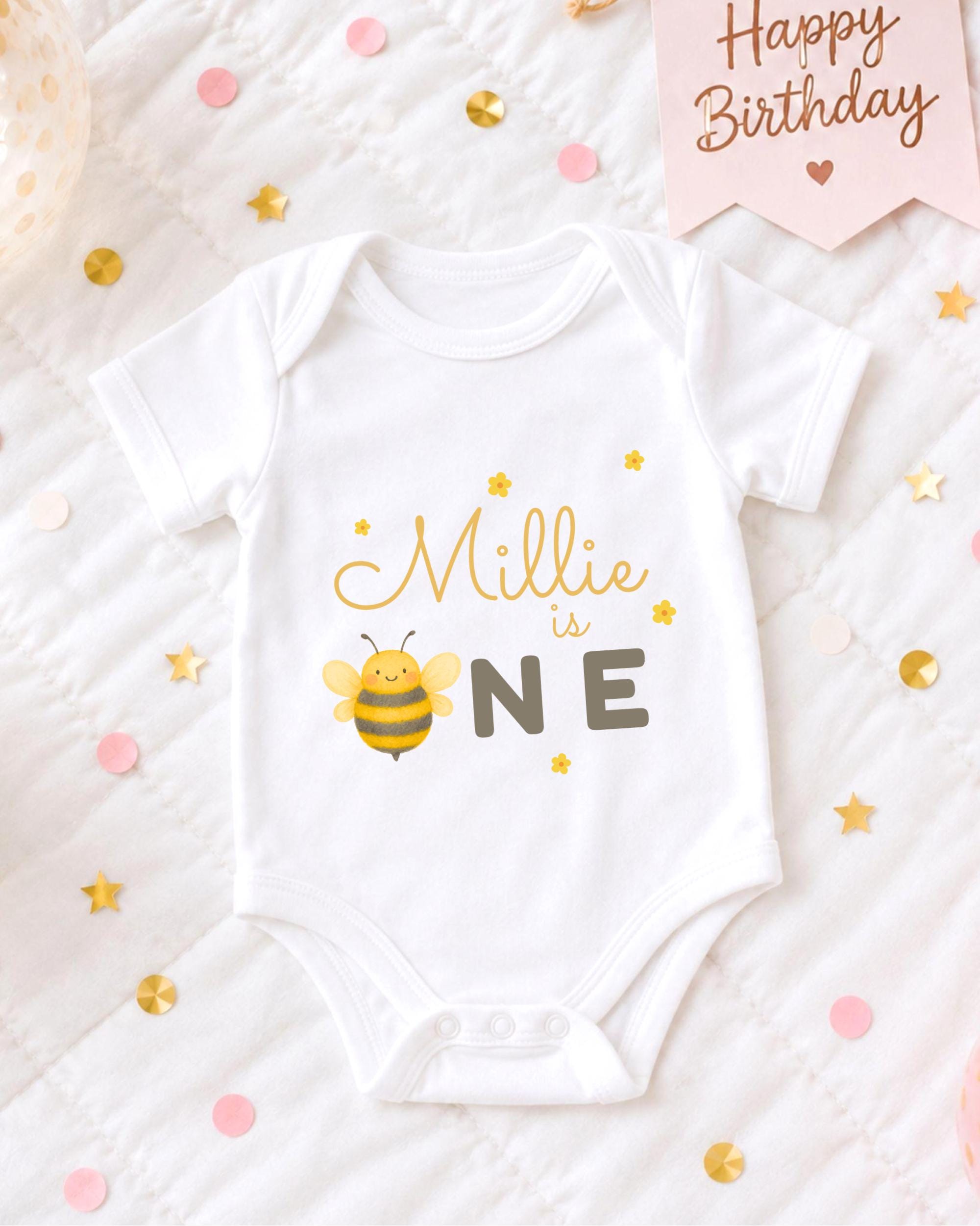 A white onesie with a bee design and the text "Milly is one" printed on it, surrounded by confetti and a birthday card.