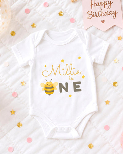 A white bodysuit with a bee design and the text "Milly is one" printed on it, surrounded by confetti and a birthday card.