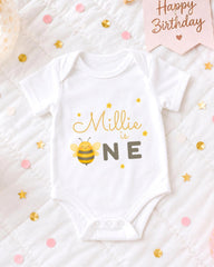 A white onesie with a bee design and the text "Milly is one" printed on it, surrounded by confetti and a birthday card.