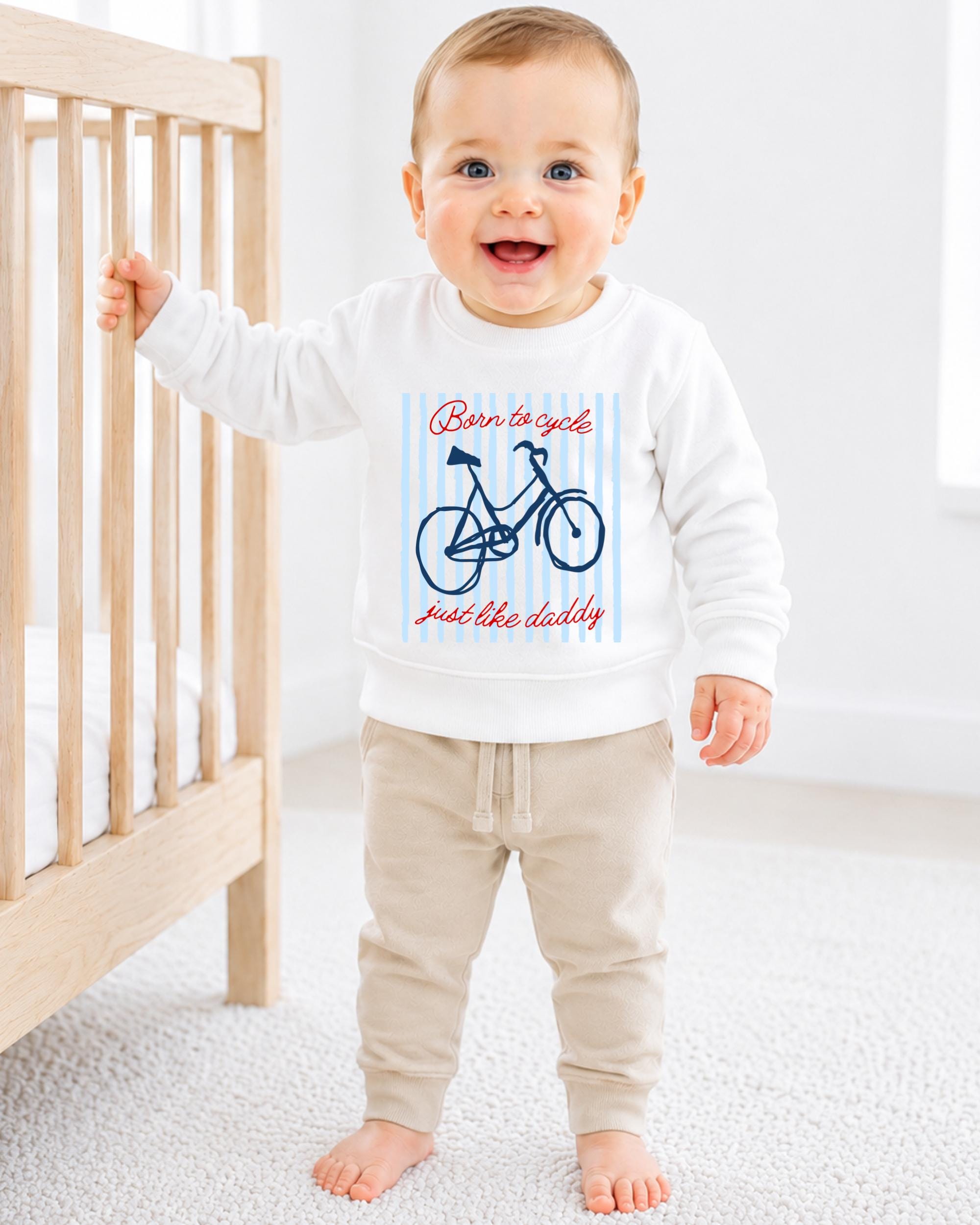 A smiling baby boy wearing a white sweatshirt with a bicycle graphic and the text "Born to cycle just like daddy" is standing next to a wooden crib.