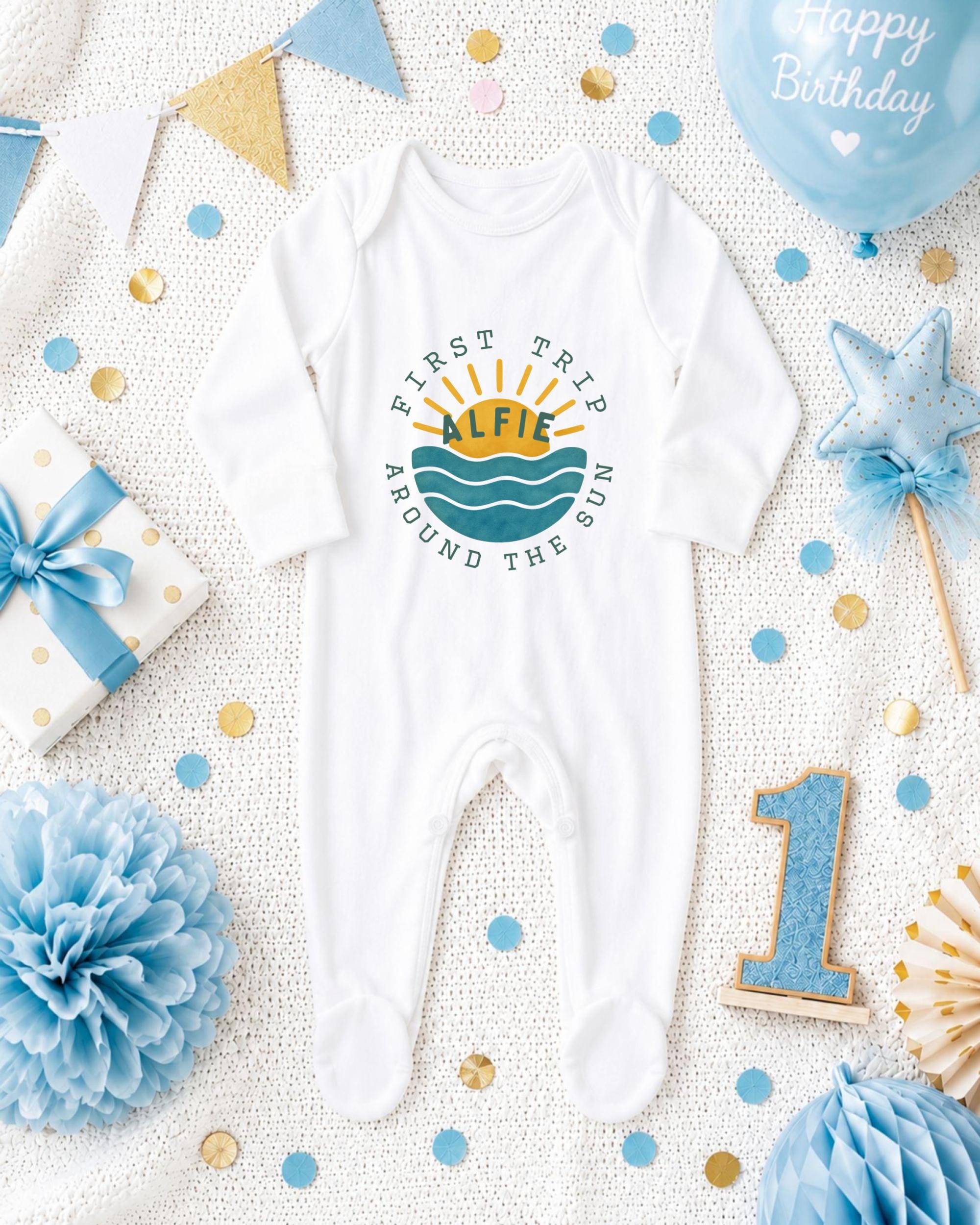 A baby onesie with the text "FIRST TRIP ALFIE AROUND THE SUN" printed on it, surrounded by various baby-related items such as a blue and gold polka dot gift box, a blue star-shaped ornament, and a blue and gold confetti-covered blanket.