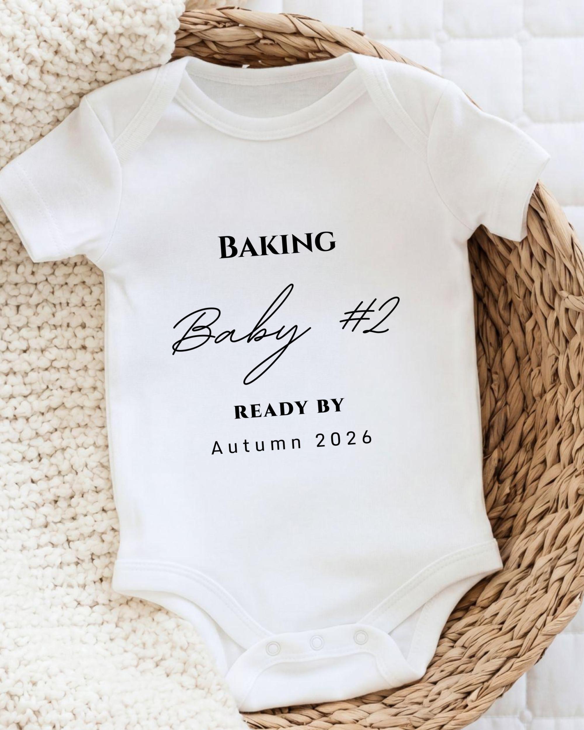 A white baby onesie with the text "BAKING Baby #2 READY BY Autumn 2026" printed on it, placed on a woven basket.