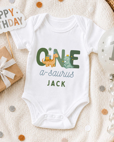 A white bodysuit with the text "ONE a-Saurus JACK" printed on it, along with illustrations of a dinosaur and a dinosaur. The bodysuit is laid out on a blanket, surrounded by a birthday card, a balloon, and confetti.