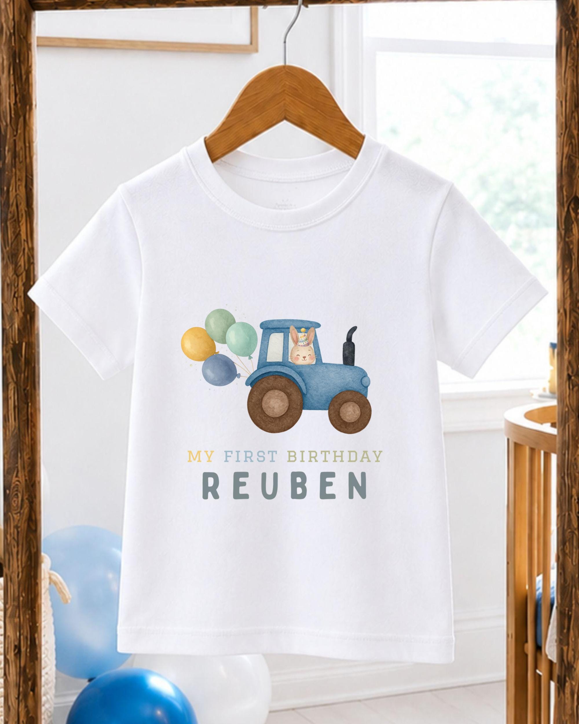 A white t-shirt with a graphic of a blue tractor carrying balloons and the text "My first birthday Reuben" printed on it.