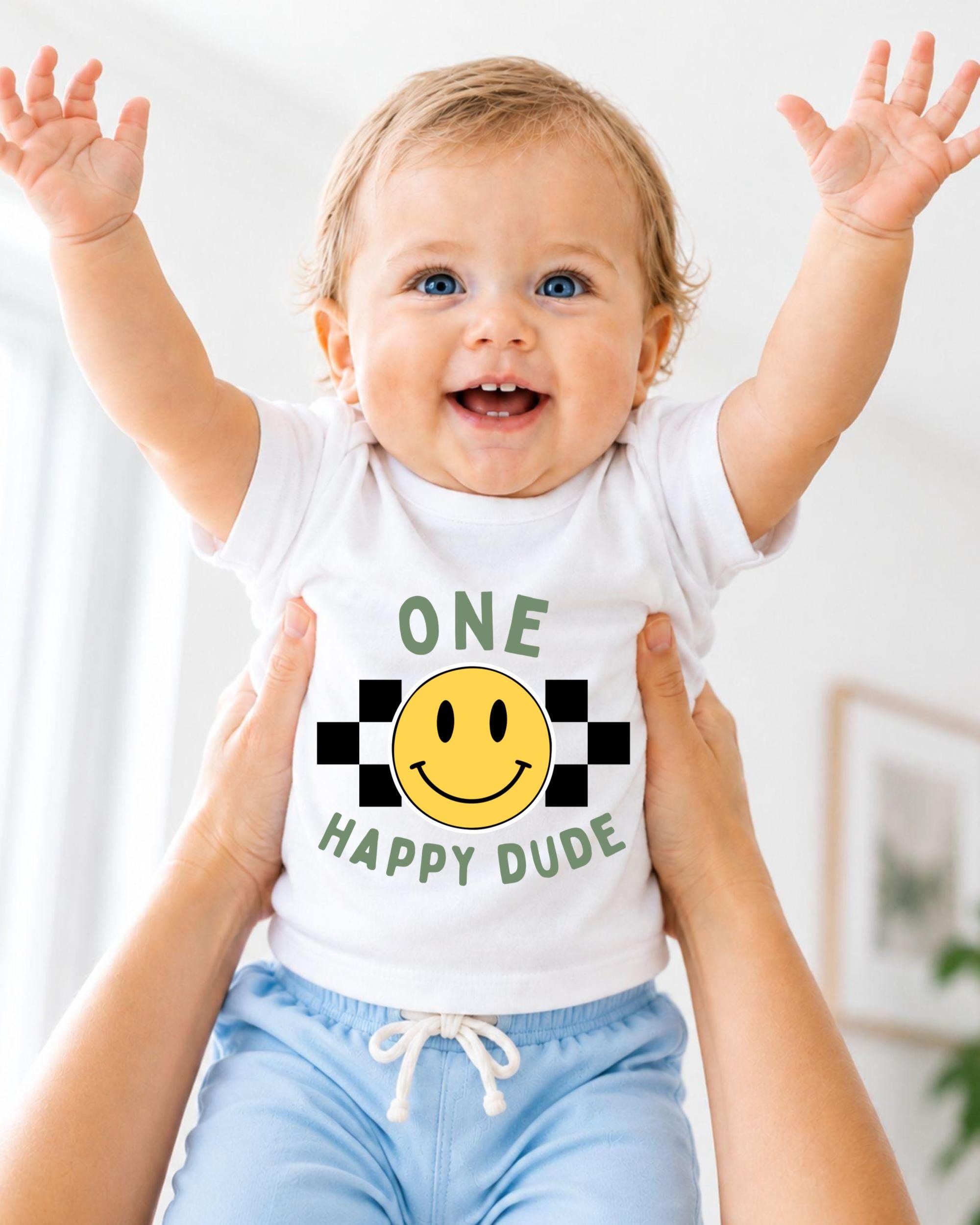 A smiling baby wearing a white shirt with a smiley face and the text "ONE HAPPY Dude" is being held up by someone's arms.