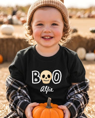 A young child wearing a black t-shirt with a skull design and the text "BOO" and "Alfie" is holding a small orange pumpkin.
