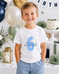 A young boy wearing a white t-shirt with the number 6 and the name "David" printed on it, smiling at the camera. The background includes birthday decorations, balloons, and a cake.