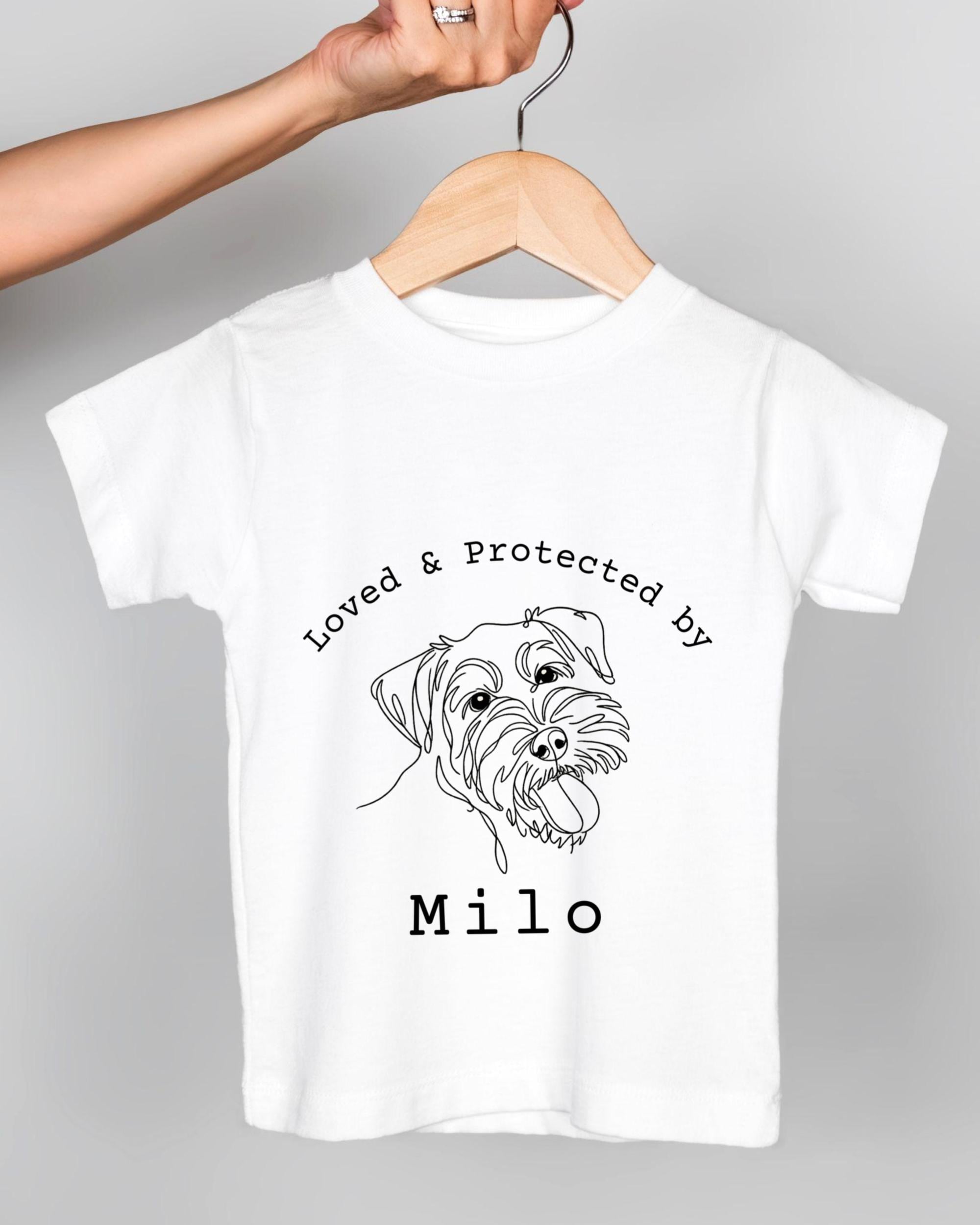 A white t-shirt with a black and white illustration of a dog's face and the text "Loved & Protected by Milo" printed on it.