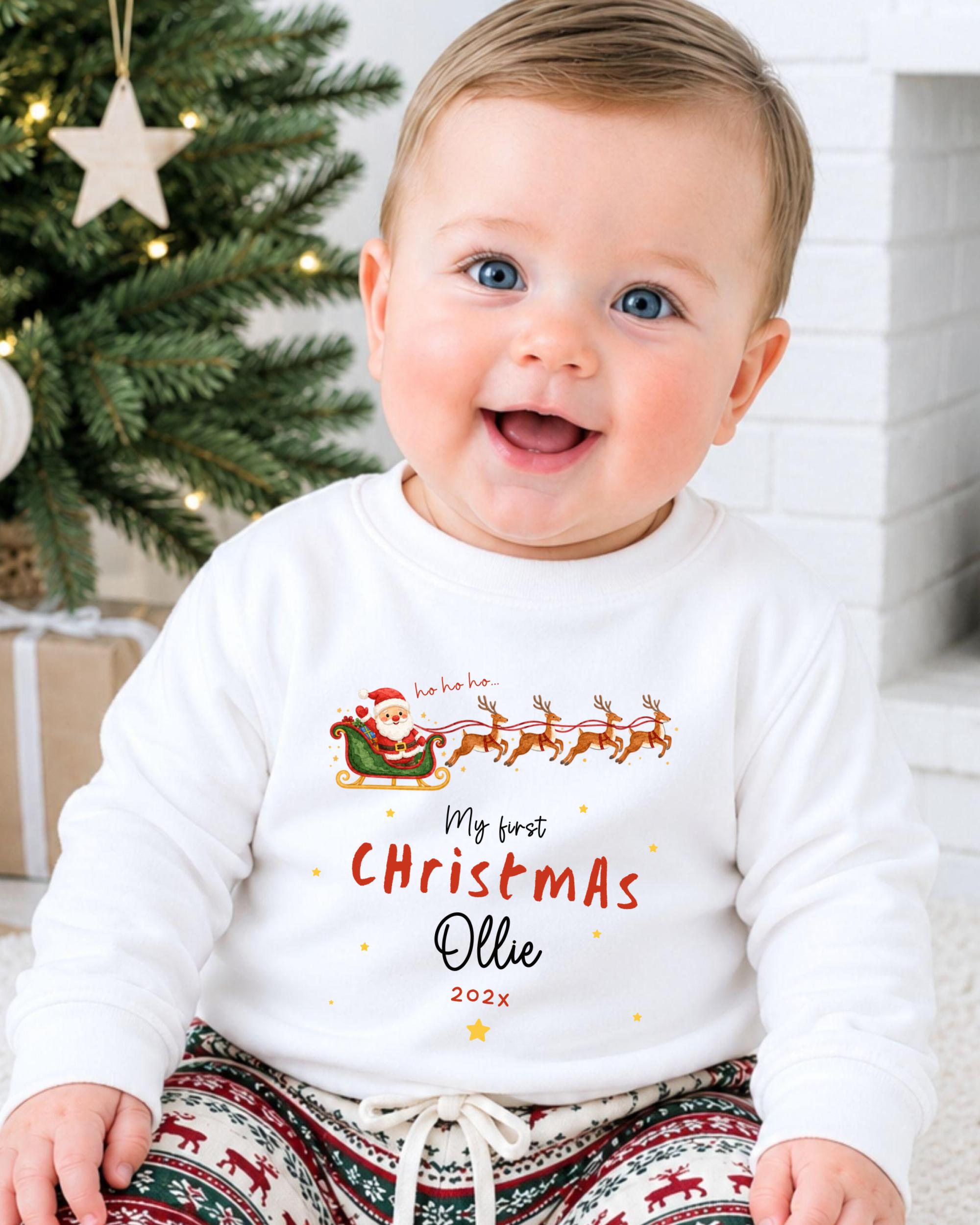 A smiling baby wearing a white shirt with a Christmas-themed design and text, sitting in front of a Christmas tree.