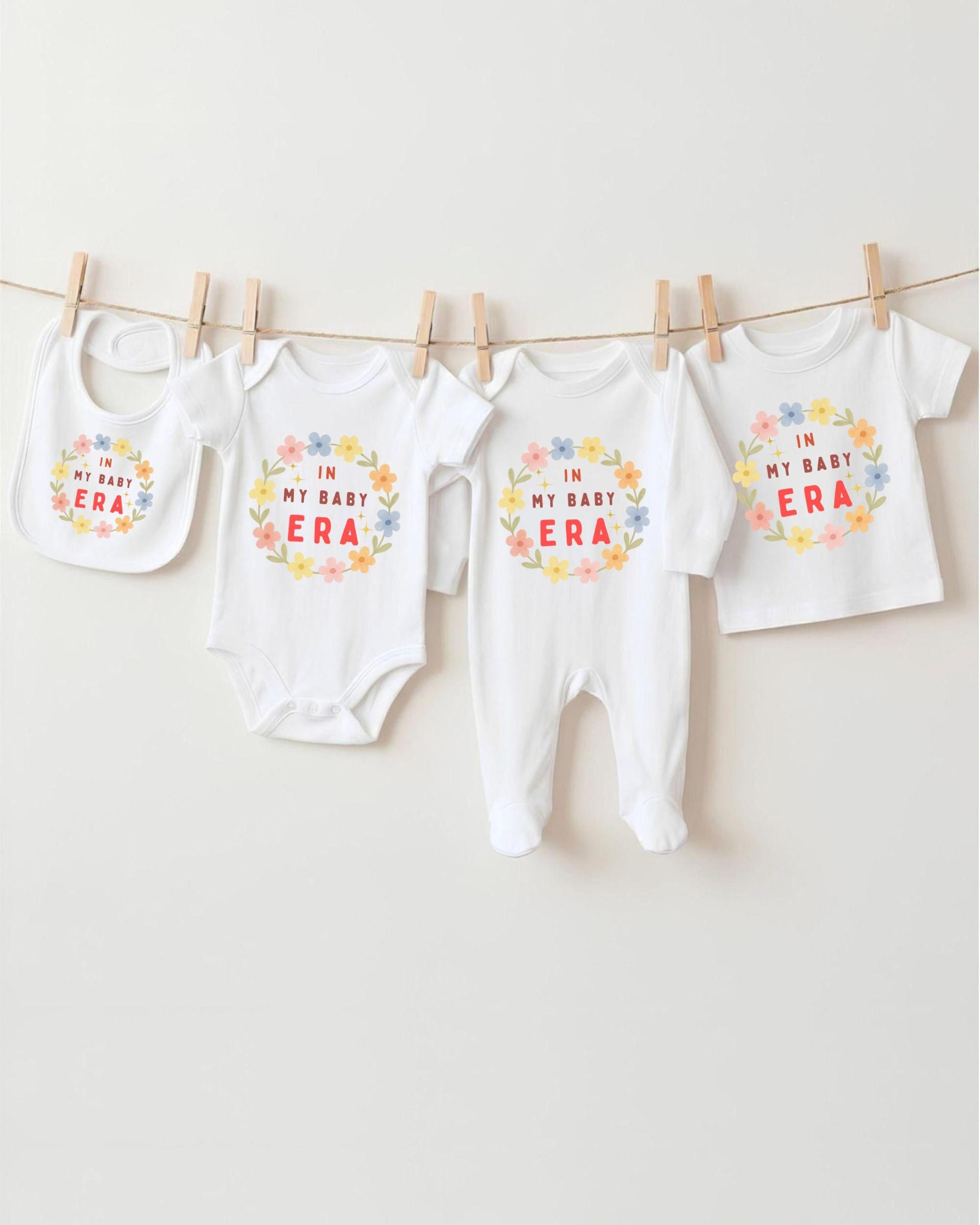 A row of four white baby clothes with the text "IN MY BABY ERA" printed on them, hanging on a clothesline.