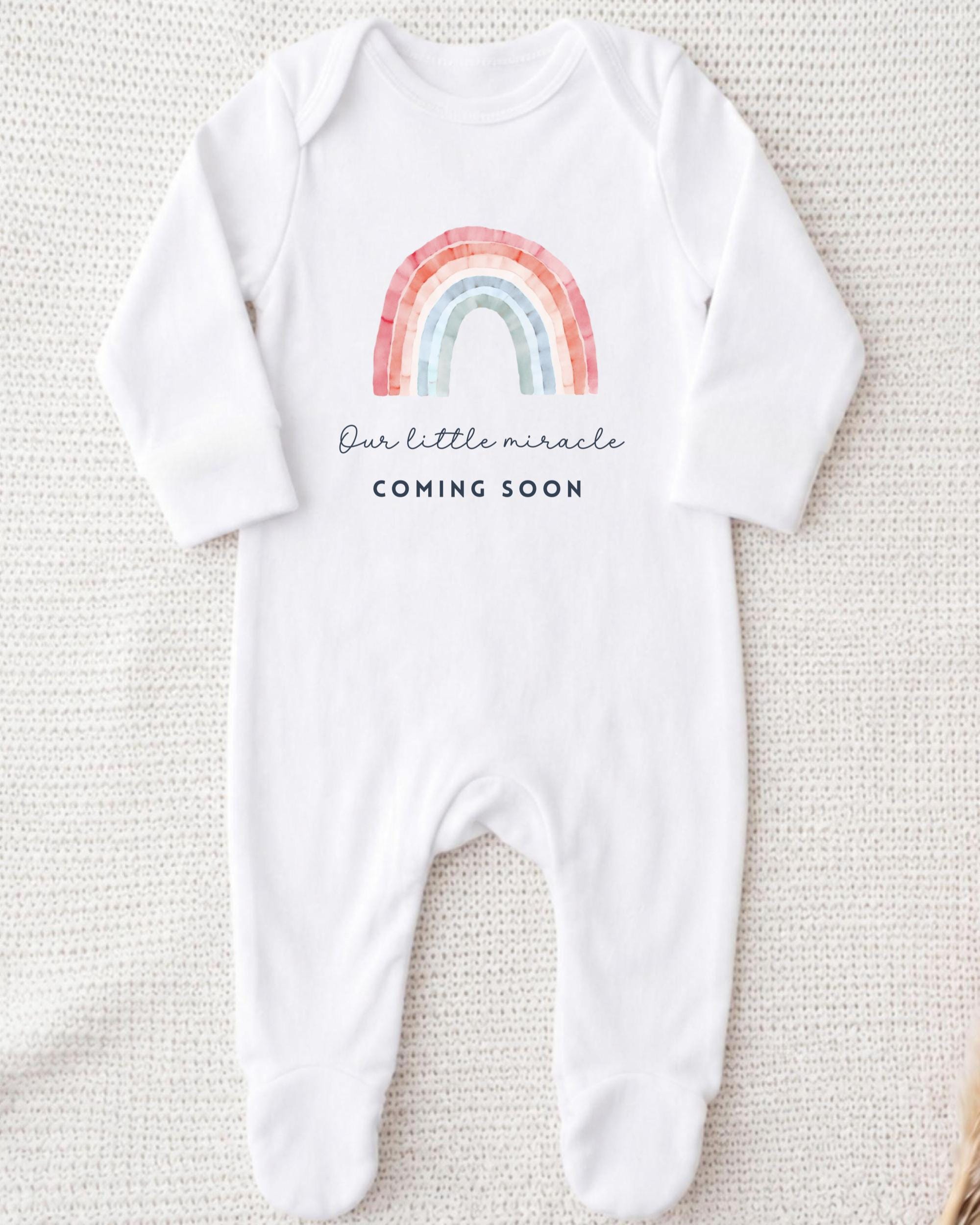 A white onesie with a rainbow graphic and the text "Our little miracle coming soon" printed on it.