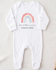 A white onesie with a rainbow graphic and the text "Our little miracle coming soon" printed on it.