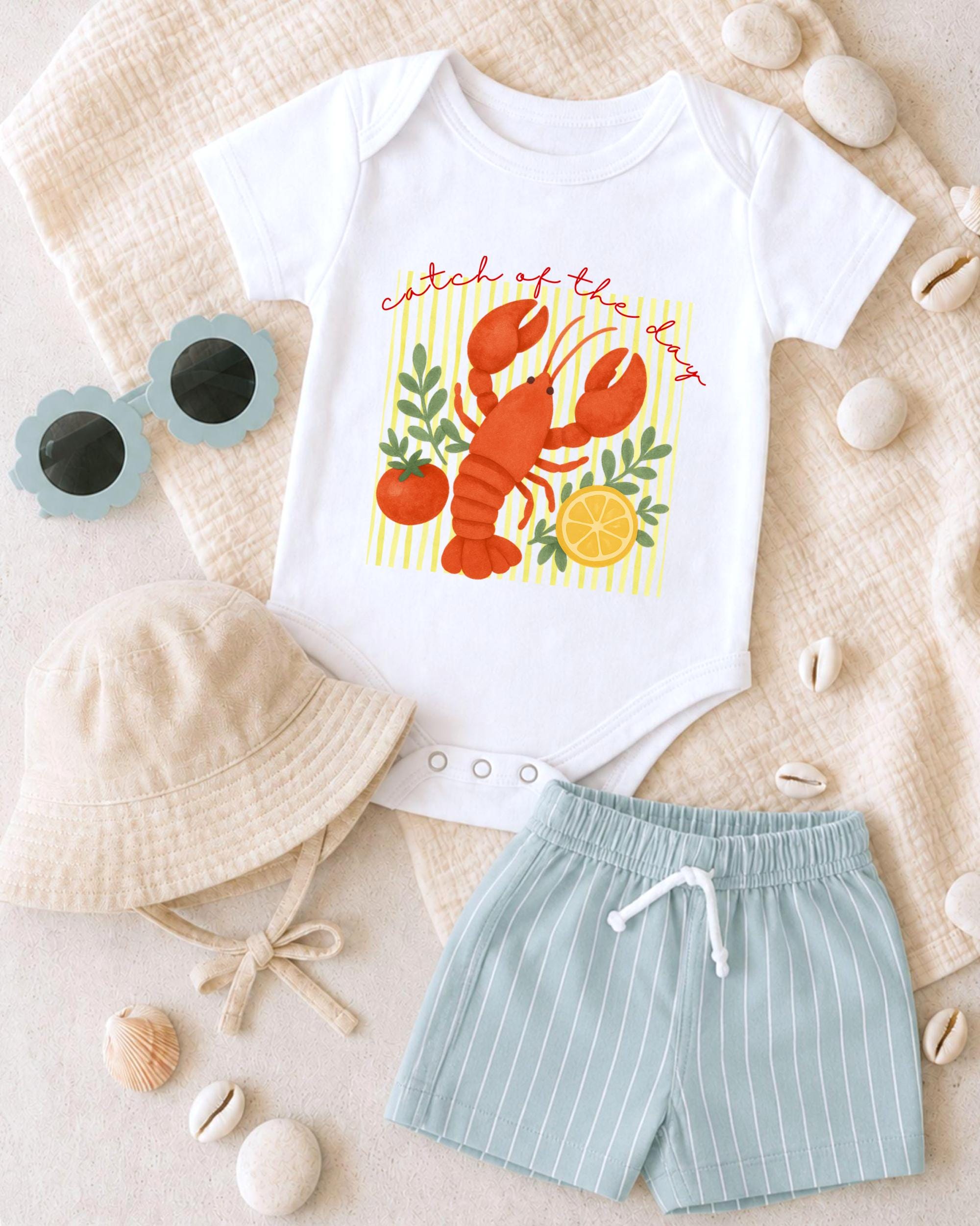A baby's outfit consisting of a white onesie with a lobster design, a beige bucket hat, and blue shorts is laid out on a beige surface, along with some accessories such as sunglasses and seashells.