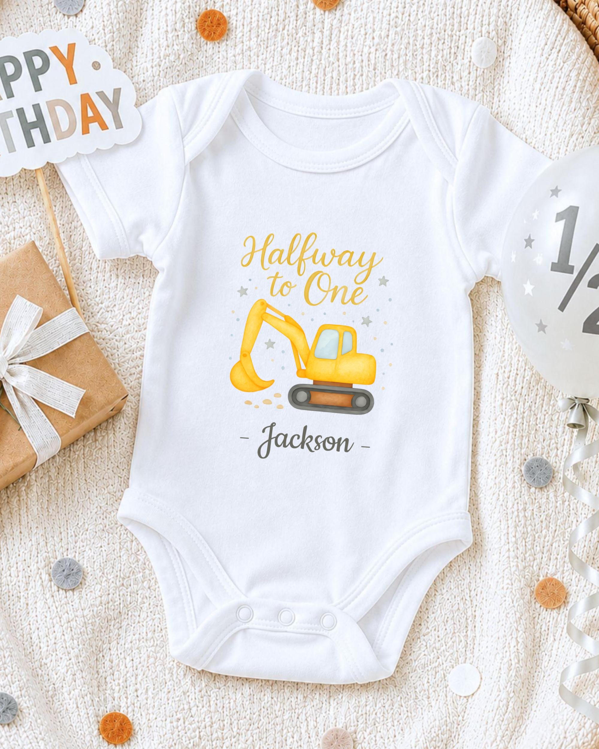 A white onesie with a yellow construction vehicle graphic and the text "Halfway to One Jackson" printed on it, along with a "Happy Birthday" sign and a gift box.
