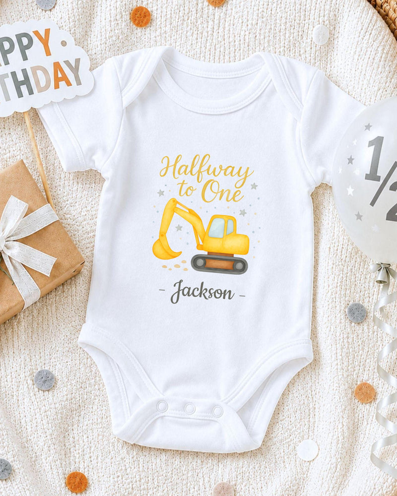 A white onesie with a yellow construction vehicle graphic and the text "Halfway to One Jackson" printed on it, along with a "Happy Birthday" sign and a gift box.