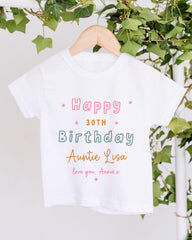 A white t-shirt with a birthday message printed on it, hanging on a wooden hanger.