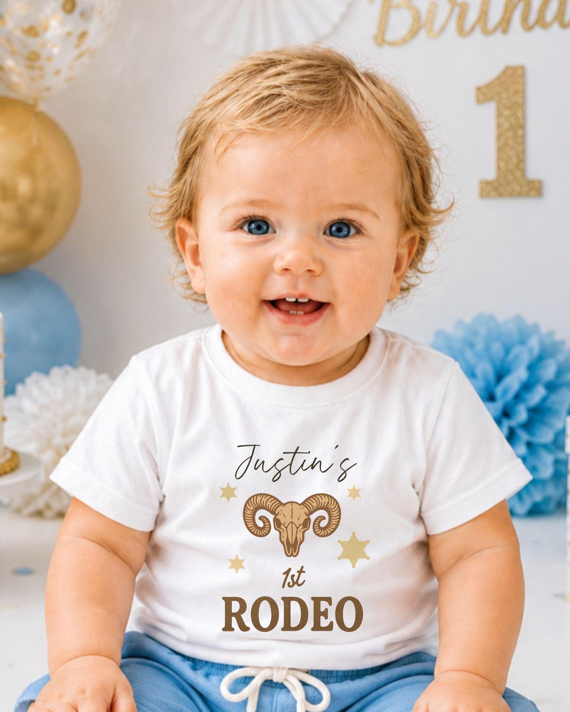 A smiling baby boy wearing a white t-shirt with a graphic of a goat and the text "Justin's 1st Rodeo" printed on it.