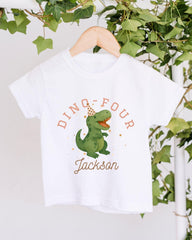 A white t-shirt with a dinosaur wearing a party hat and the text "DINO FOUR JACKSON" printed on it, hanging on a wooden hanger.