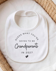 A white bib with text that says "Guess what you're going to be Grandparents in 2027" printed on it.