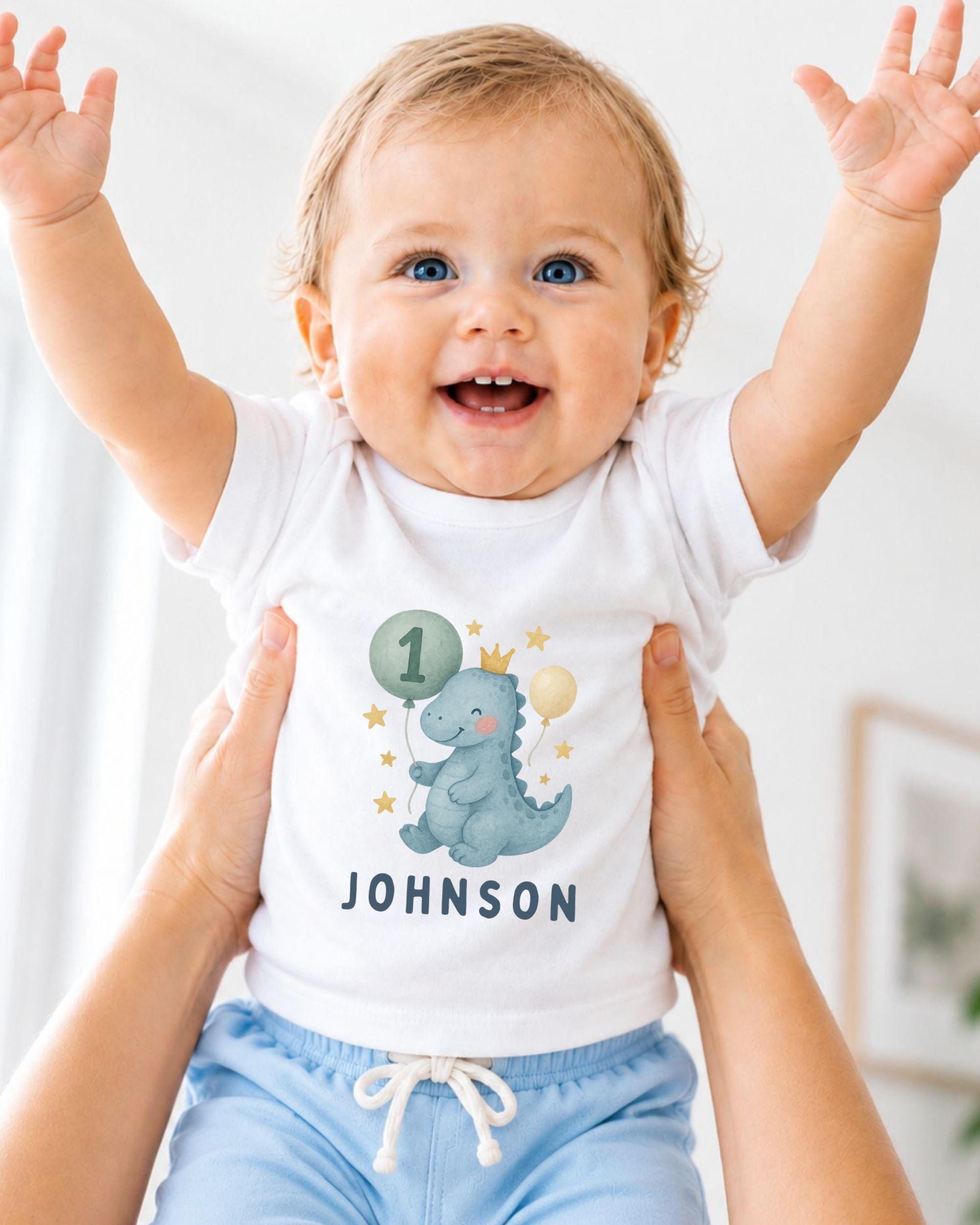 A smiling baby wearing a white t-shirt with a dinosaur graphic and the name "Johnson" printed on it, being held up by someone's arms.