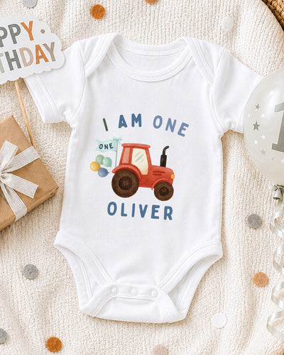 A white bodysuit with a red tractor design and the text "I AM ONE OLIVER" printed on it, along with a birthday sign and a balloon.