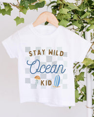 A white t-shirt with a graphic design featuring a checkered pattern, a jellyfish, and a surfboard, along with the text "STAY WILD Ocean KID".