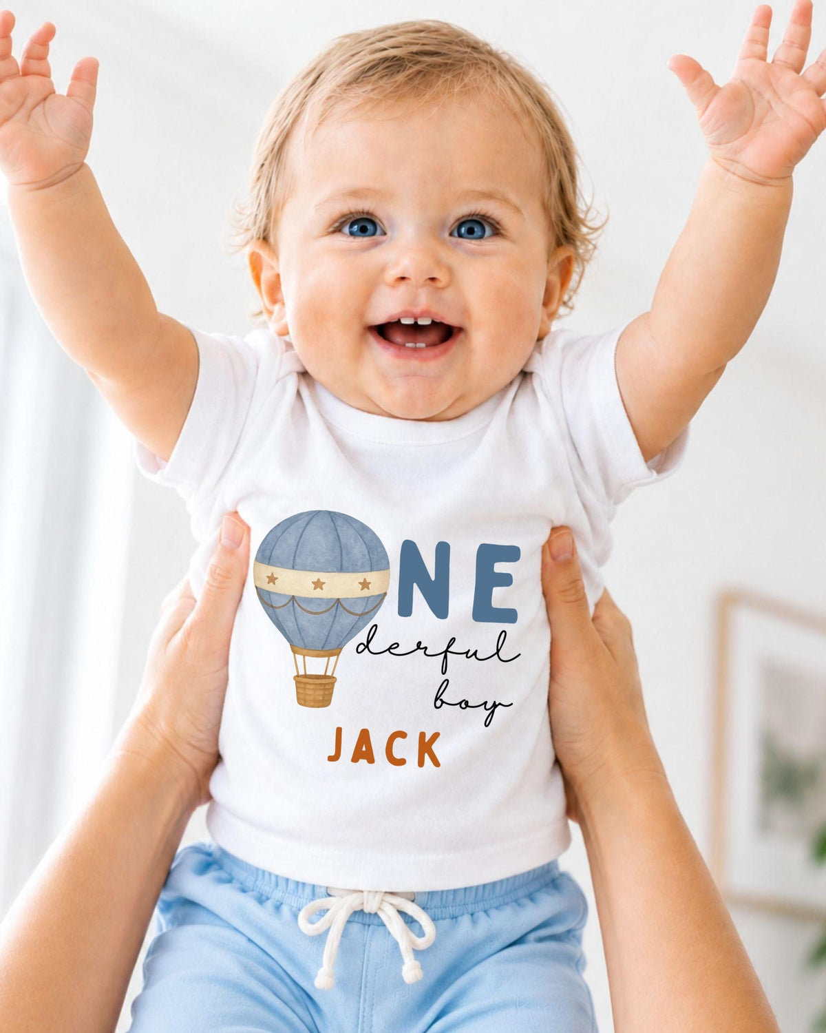 A smiling baby wearing a white shirt with a hot air balloon and the text "One derful boy Jack" is being held up by someone's arms.