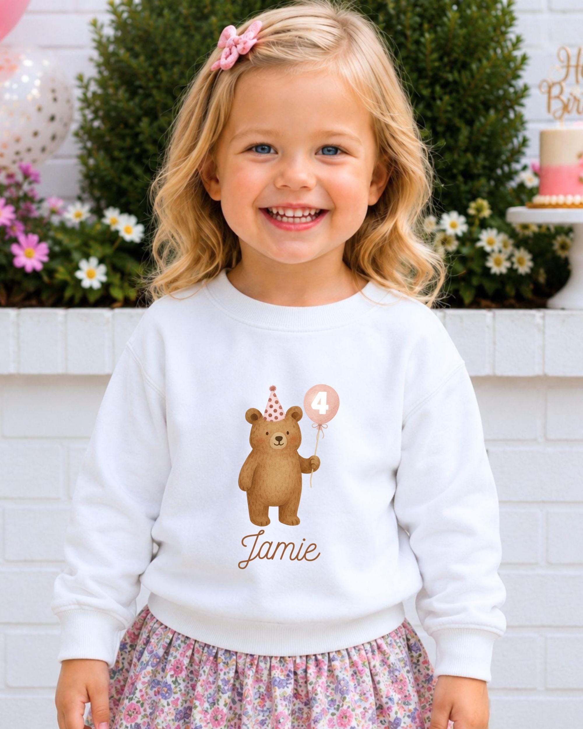 A young girl wearing a white sweater with a teddy bear and a birthday balloon, smiling at the camera.