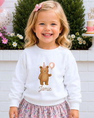 A young girl wearing a white sweater with a teddy bear and a birthday balloon, smiling at the camera.